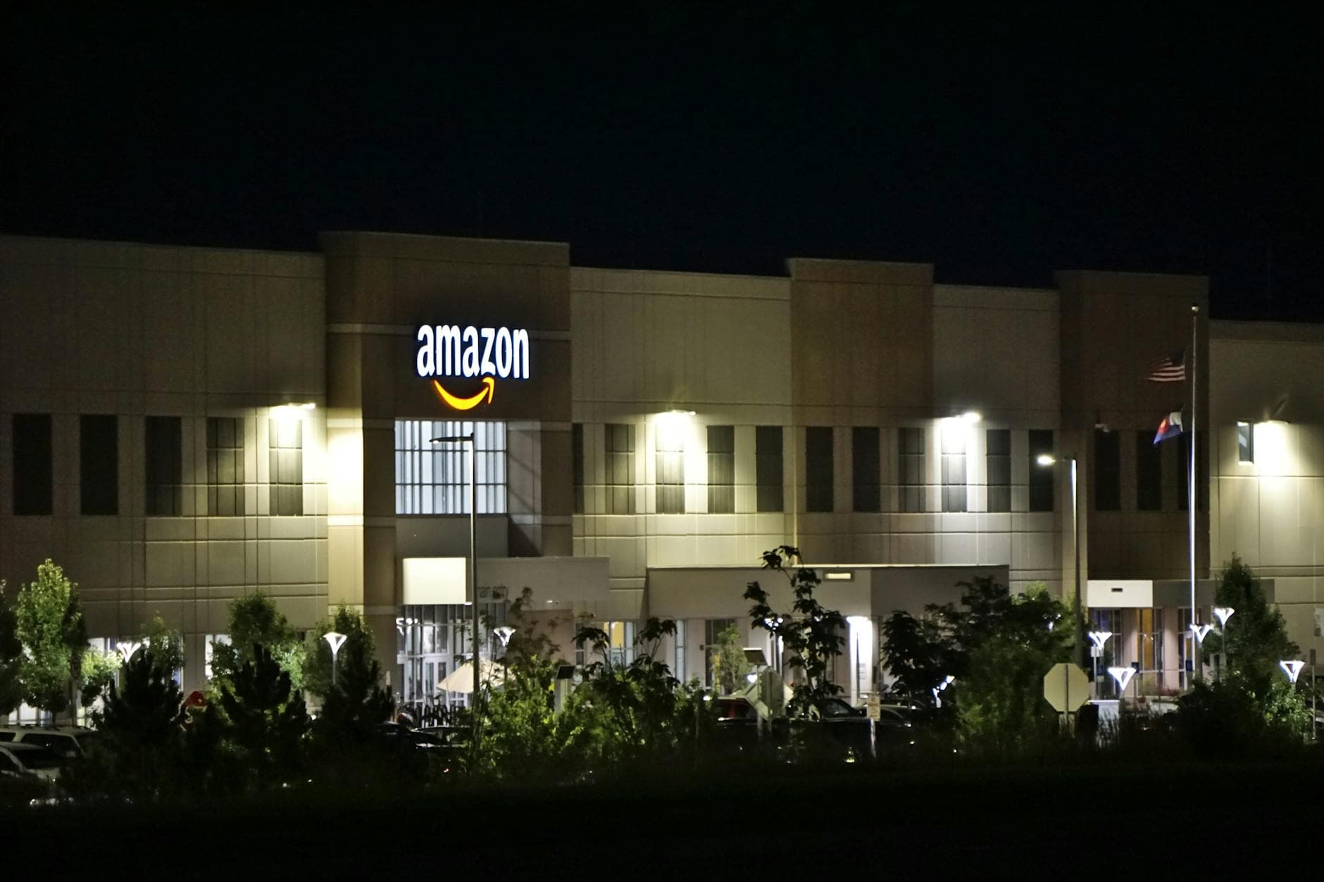 Amazon warehouse building illuminated at night with trees and signage.