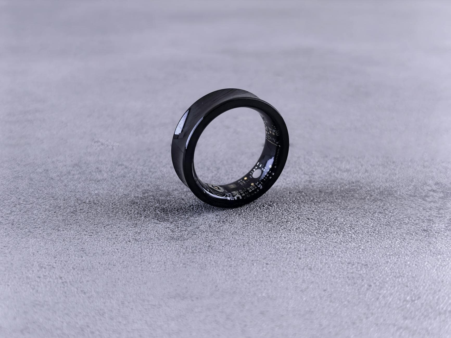 A modern black smart ring with visible circuitry resting on a textured concrete surface.